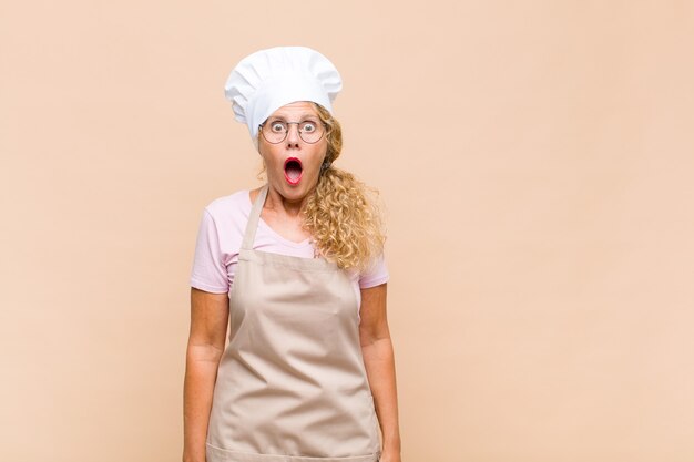 Middle aged woman baker feeling terrified and shocked, with mouth wide open in surprise
