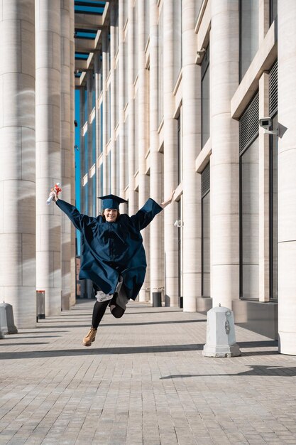 Middle aged woman in blue graduation gown and cap jumping in\
campus with pillars