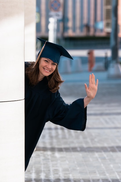 Middle aged woman in blue graduation gown and cap peeking\
around pillars in campus