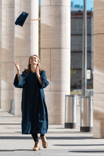 Middle aged woman in blue graduation gown throwing her cap in\
campus with pillars