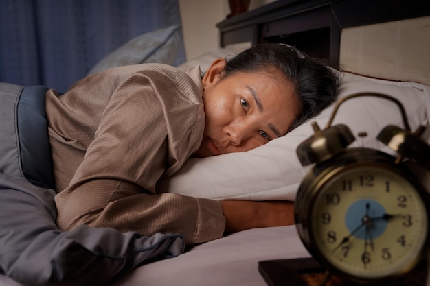 Middle aged woman depressed and stressed lying in bed looking\
at the clock from insomnia