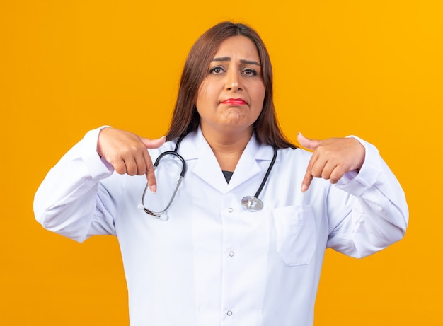 Middle aged woman doctor in white coat with stethoscope looking at front with sad expression pointing at herself standing over orange wall