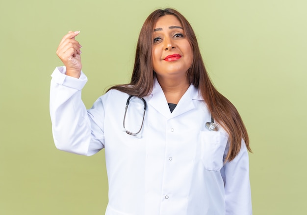 Middle aged woman doctor in white coat with stethoscope looking at front with smile on face making money gesture rubbing fingers standing over green wall