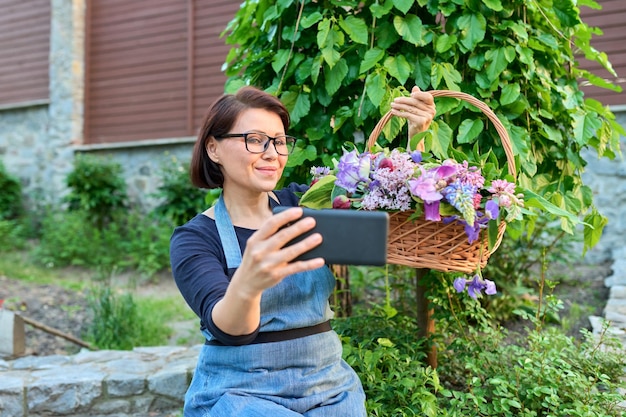 Middle aged woman in garden with basket of fresh spring flowers taking selfie photo