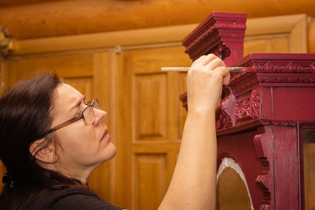 Middle aged woman in glasses painting patterned antique cupboard in red color with little brush Home workshop for renovation of furniture New life for old things