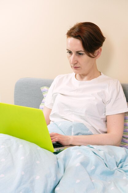 Middle-aged woman lies in bed and works on a laptop. A workaholic woman checks her email before bed. Stress and overwork from challenging tasks at work.