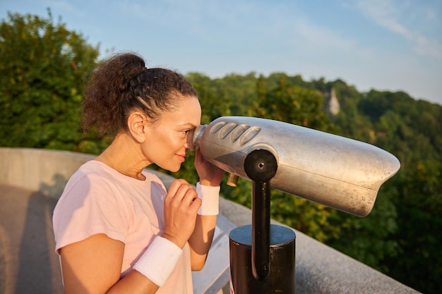 Middle aged woman looks through a telescope at the city. Athletic woman in a pink t-shirt is watching the beautiful green nature and the river background through stationary city binoculars