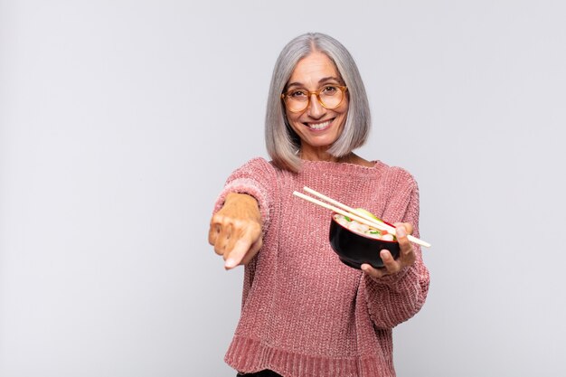 Middle aged woman pointing at camera with a satisfied, confident, friendly smile, choosing you asian food concept