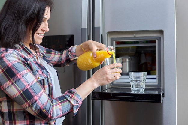 Женщина средних лет наливает апельсиновый сок в стакан из холодильника