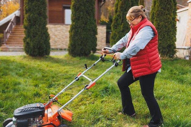 Middle aged woman in red vest using lawn mower on backyard
looking at camera female gardener working in summer or autumn
cutting grass in backyard concept of gardening work nature