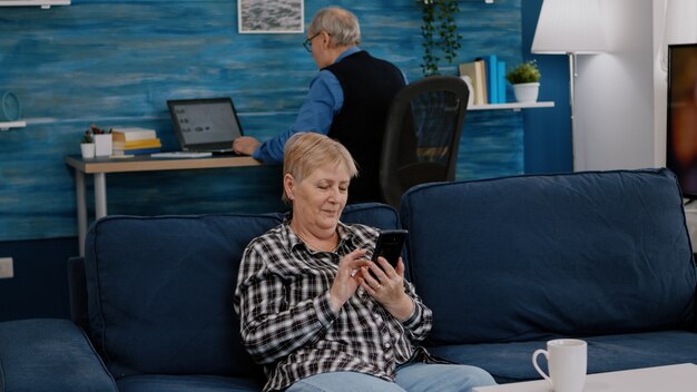 Middle aged woman relaxing holding smartphone reading e book sitting on sofa at home while senior ad...