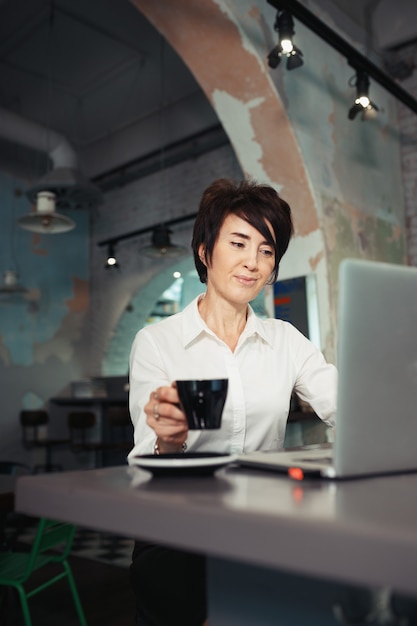 Una donna di mezza età si siede in un bar, beve caffè e lavora al computer