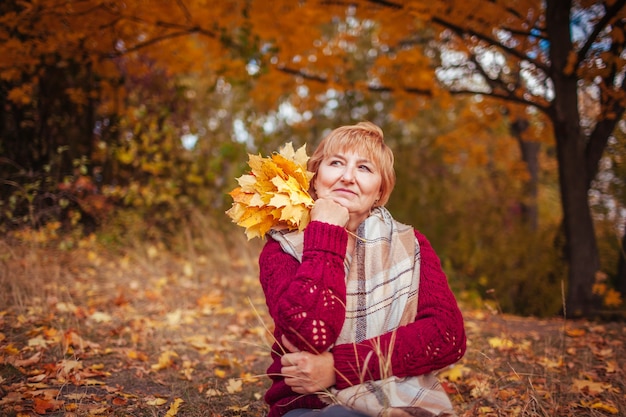 Middle-aged woman surrounded with yellow trees relaxes in autumn forest. Lady enjoying nature holding bouquet of leaves