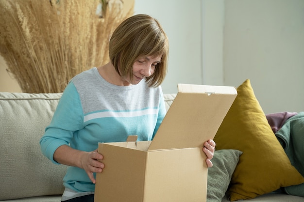 Middle aged woman unpacking cardboard box sitting on sofa at home