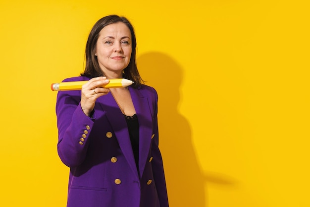 Middle aged woman wearing purple blazer is holding giant pencil on yellow background