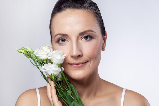 Middle aged woman with clean healthy skin holding flowers in hadns looking at camera with cute smile