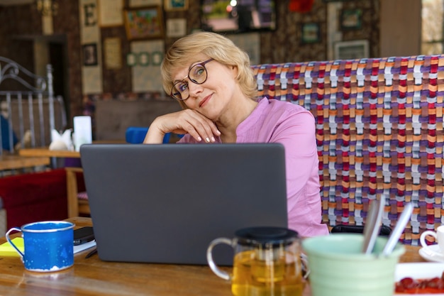 A middle-aged woman with a laptop works in a cafe in the office, she is a freelancer. Woman in glasses sitting at the table with a Cup of herbal tea. She's in a good mood.