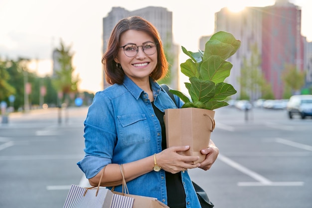 Middle aged woman with paper shopping bags with buying plant looking at camera