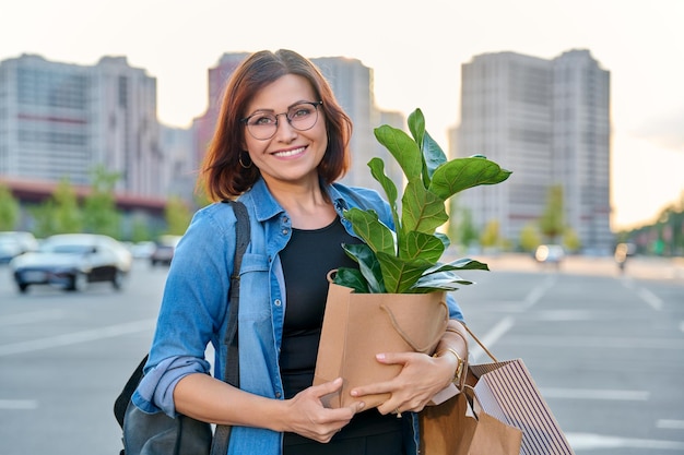 Middle aged woman with paper shopping bags with buying plant looking at camera