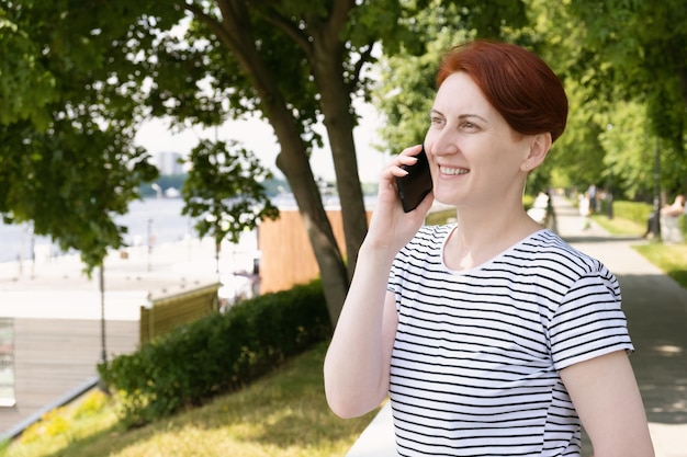 Foto una donna di mezza età con i capelli rossi si trova sull'argine del parco cittadino sorridendo mentre parla al telefono