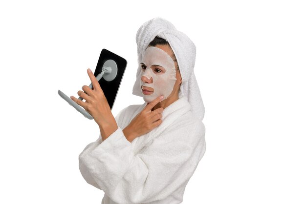 Middle-aged woman with wet sheet mask on her face looks in the mirror, wearing a white towel on her head, white isolated background.