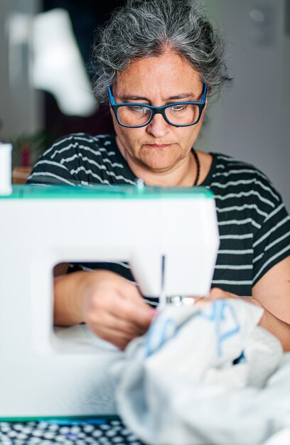Женщина средних лет с белыми волосами шьет на швейной машинке дома.