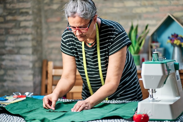 middle-aged woman with white hair, wearing glasses and sewing tape around her neck, working at the sewing table at home.