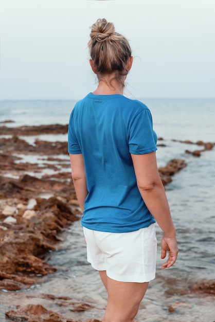 日没時に海沿いの崖の上に立っている髪のお団子と青いTシャツの中年女性。バックシュート、Tシャツのモックアップ