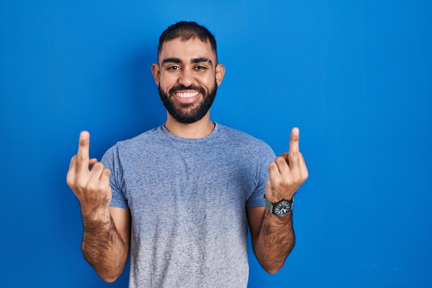 Мужчина с Ближнего Востока с бородой, стоящий на синем фоне, показывает средний палец, провоцирует тебя на плохое выражение лица и грубо кричит от возбуждения