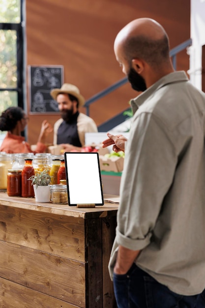 Foto uomo del medio oriente che guarda un tablet elettronico con uno schermo bianco vuoto posto su un tavolo pieno di prodotti alla rinfusa in contenitori di vetro riutilizzabili.