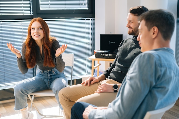 Middle shot portrait of cheerful young redhead businesswoman talking and discussing new ideas with creative business team, during brainstorming of start-up projects in modern office room near window.