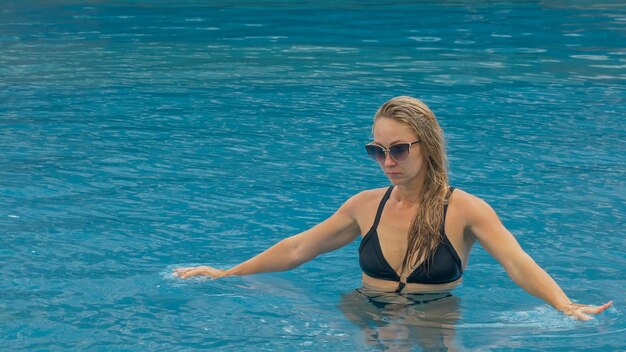 Middle shot pretty woman drinking blue cocktail alcohol liquor in swimming pool at hotel Portrait of sexy girl in pool outdoor Beautiful caucasian women with blond hair black swimsuit sunglasses