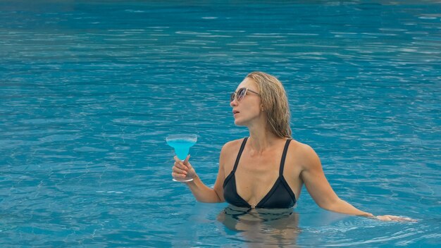 Middle shot pretty woman drinking blue cocktail alcohol liquor in swimming pool at hotel Portrait of sexy girl in pool outdoor Beautiful caucasian women with blond hair black swimsuit sunglasses