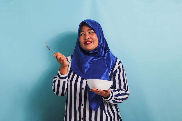 A middleaged Asian woman in hijab is holding a white bowl and spoon isolated on a blue background