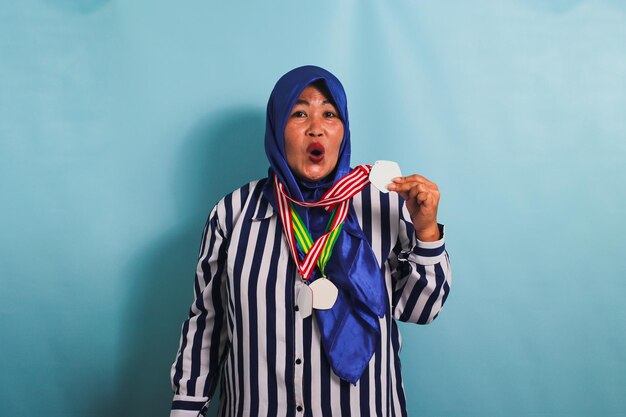 ヒジャブを着た中年アジア人女性が青い背景で孤立している間メダルを展示しています