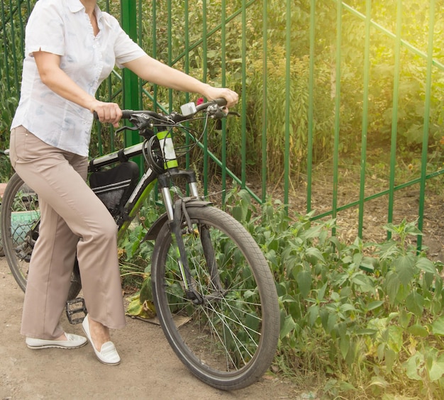 軽いズボンとシャツを着た中年の魅力的なスリムな女性が公園の自転車の近くに立つ