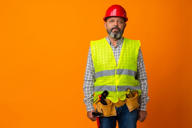 Фото Бородатый мужчина средних лет-строитель в униформе и каске с инструментами на оранжевом фоне