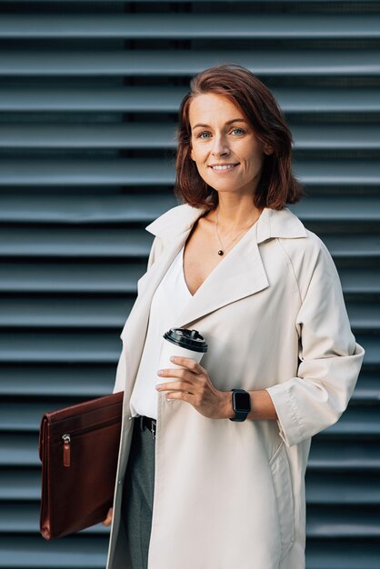 중년 사업가 여성 은 야외 에 서 있는 동안 커피 컵 과 가죽 폴더 를 들고 있다
