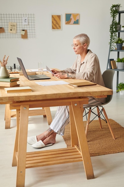 Middleaged businesswoman in slippers sitting at table in home office and reading contract content du