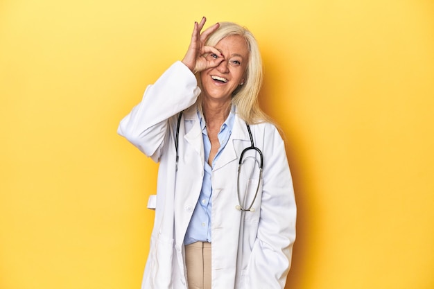 Photo middleaged caucasian female doctor on yellow studio backdropexcited keeping ok gesture on eye