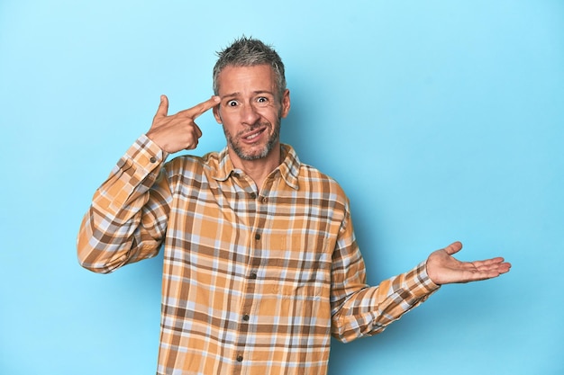 Middleaged caucasian man on blue backdrop holding and showing a product on hand