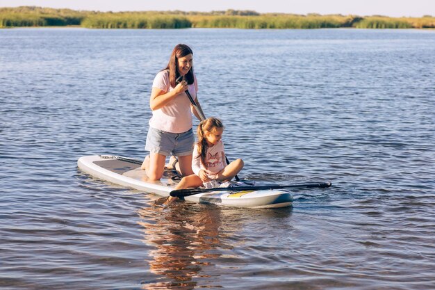 Middleaged Caucasian mother with little daughter sup boarding both smiling and looking down at water Active lifestyle and family time together Teaching children to do sports