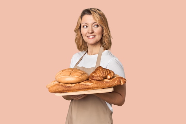 Middleaged Caucasian woman baker with bread tray looks aside smiling cheerful and pleasant