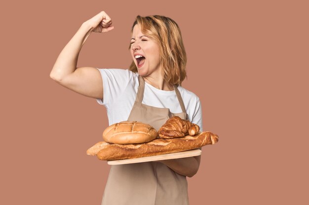 Middleaged Caucasian woman baker with bread tray raising fist after a victory winner concept