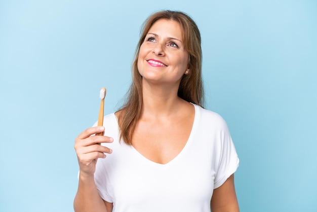 Middleaged caucasian woman brushing teeth isolated on blue background looking up while smiling