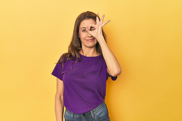 Middleaged caucasian woman on yellow excited keeping ok gesture on eye