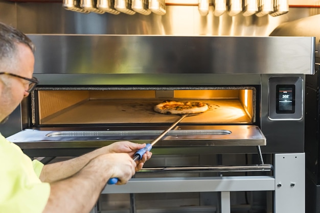 Foto cuoco di mezza età che tira fuori la pizza cotta da una cucina di panetteria a forno elettrico