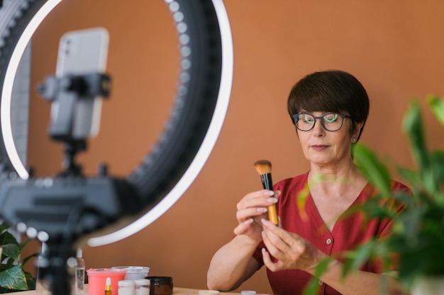 Blogger di bellezza femminile di mezza età o influencer con prodotti per il trucco che registrano video nella stanza di casa utilizzando lampada ad anello e smartphone