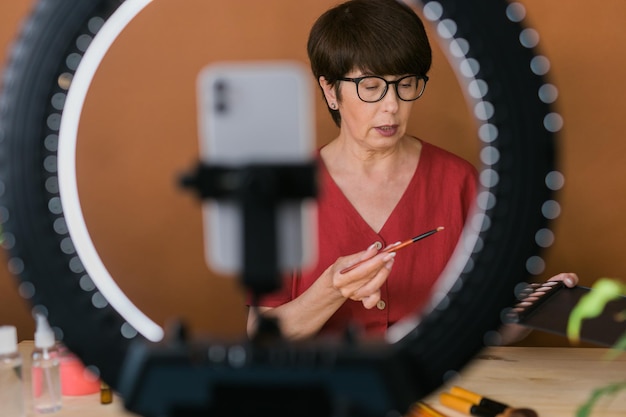 사진 중년 여성 뷰티 블로거 또는 메이크업 제품이 있는 인플루언서, 집에서 링 램프와 스마트폰을 사용하여 비디오 녹화