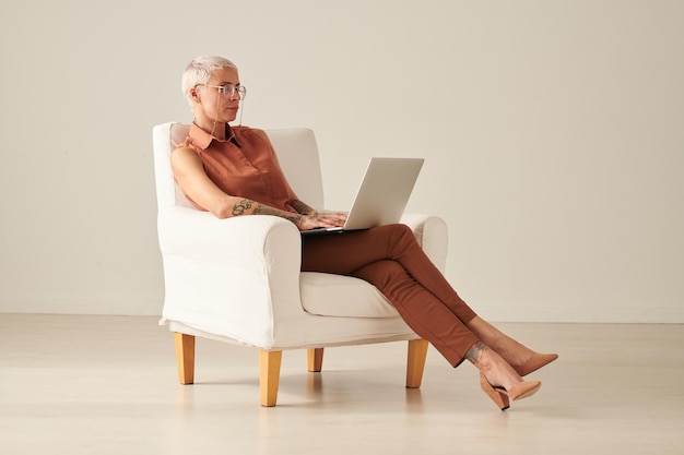 Middleaged freelance woman in stylish outfit sitting into comfortable armchair and using laptop whil